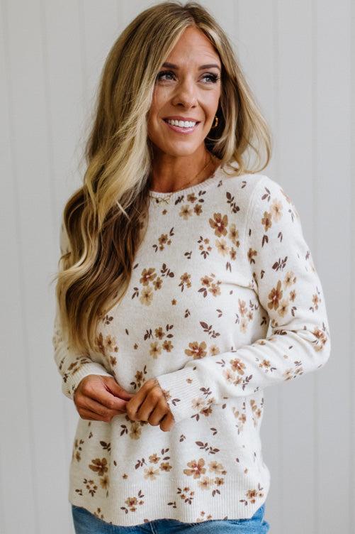 A woman with long blonde hair wearing a cream-colored knit sweater with a brown and tan floral pattern, paired with blue jeans, standing against a white paneled wall.