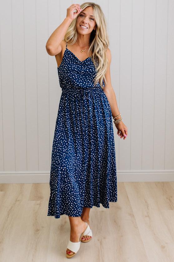 Woman wearing a navy blue dress with white polka dots, standing against a light wooden paneled wall.