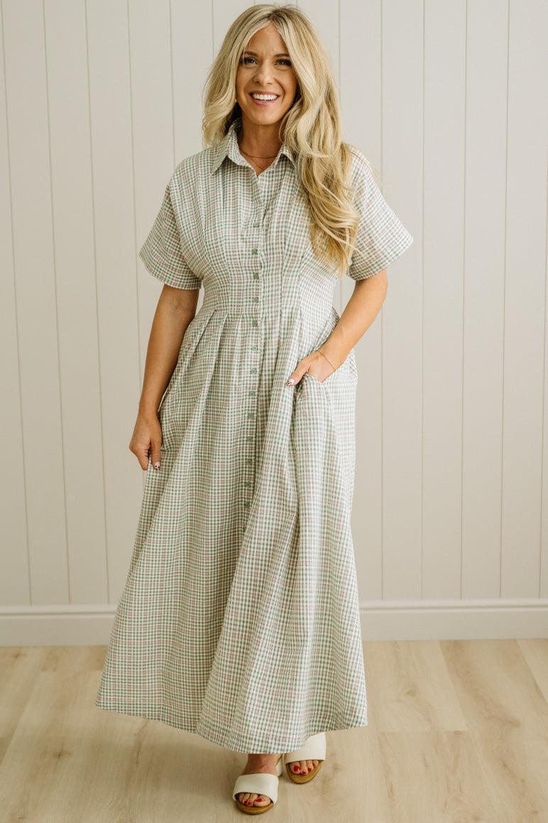 Woman wearing a checkered dress standing against a light wooden paneled wall.