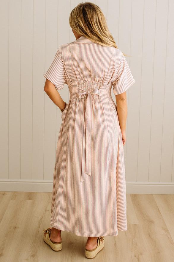 Woman wearing a long pink and white striped dress with a belt, standing against a light wooden paneled wall.