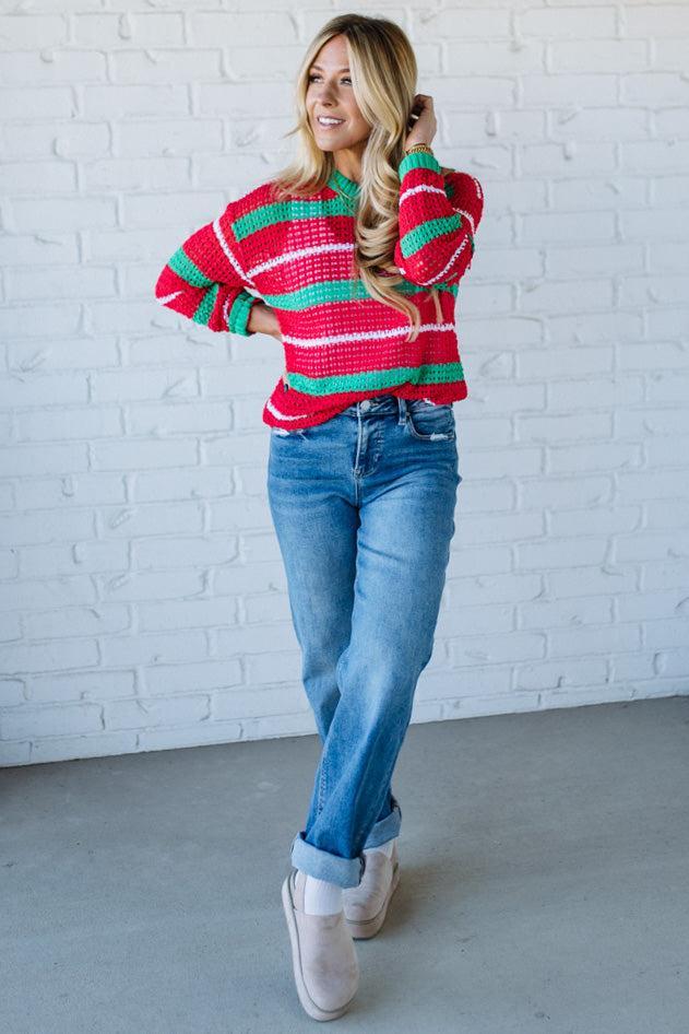 Woman wearing a red and green striped sweater and blue jeans against a white brick wall.
