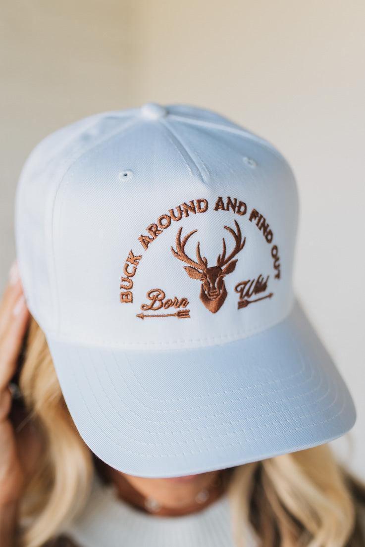 white cap with brown deer emblem and text held by a person against a neutral background