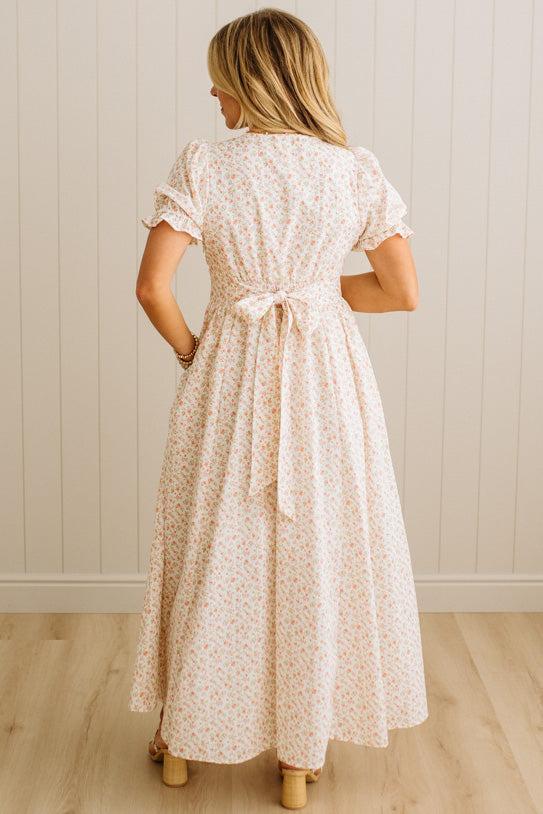 Woman wearing a white floral dress with a belt, standing against a light wooden paneled wall.