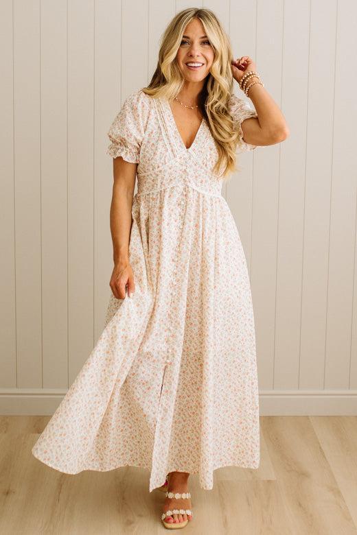 Woman wearing a white floral dress against a light wooden paneled wall.