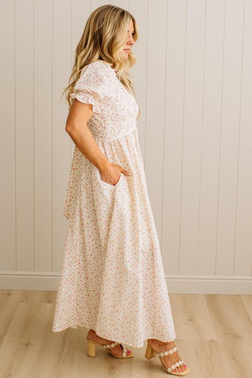 Woman wearing a white floral dress standing against a light wooden paneled wall.