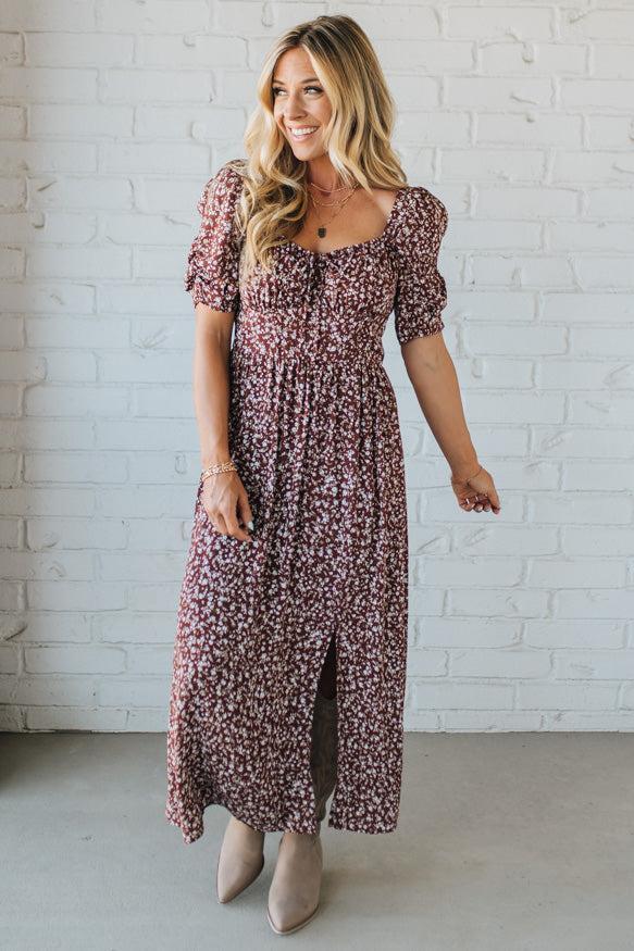 Woman wearing a floral dress standing against a white brick wall.