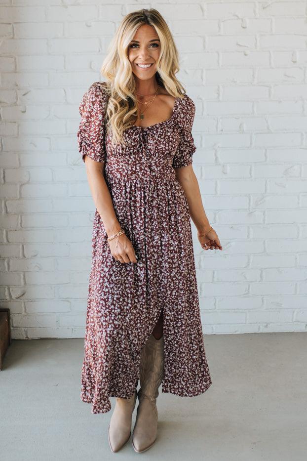 Woman wearing a floral dress standing against a white brick wall.