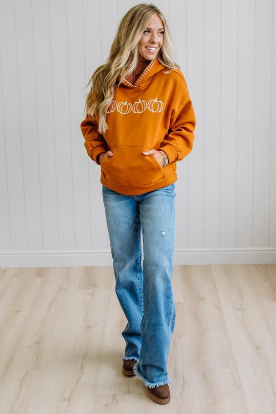 A woman wearing a burnt orange hoodie with a white pumpkin graphic on the chest, paired with light-wash blue jeans and brown shoes, standing in a room with white paneled walls and light wood floors.