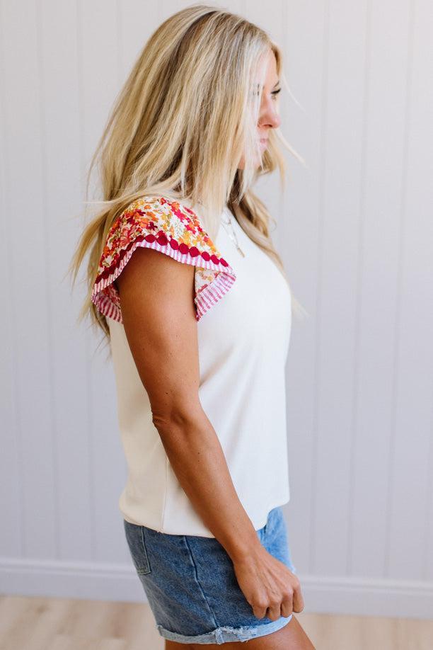 Woman wearing a white top with colorful ruffled sleeves and denim shorts against a plain background
