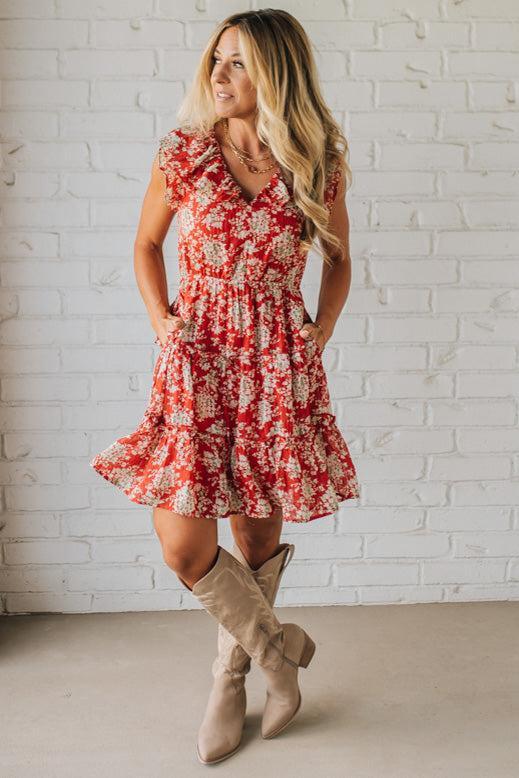 Woman wearing a red floral dress and beige boots against a white brick wall.