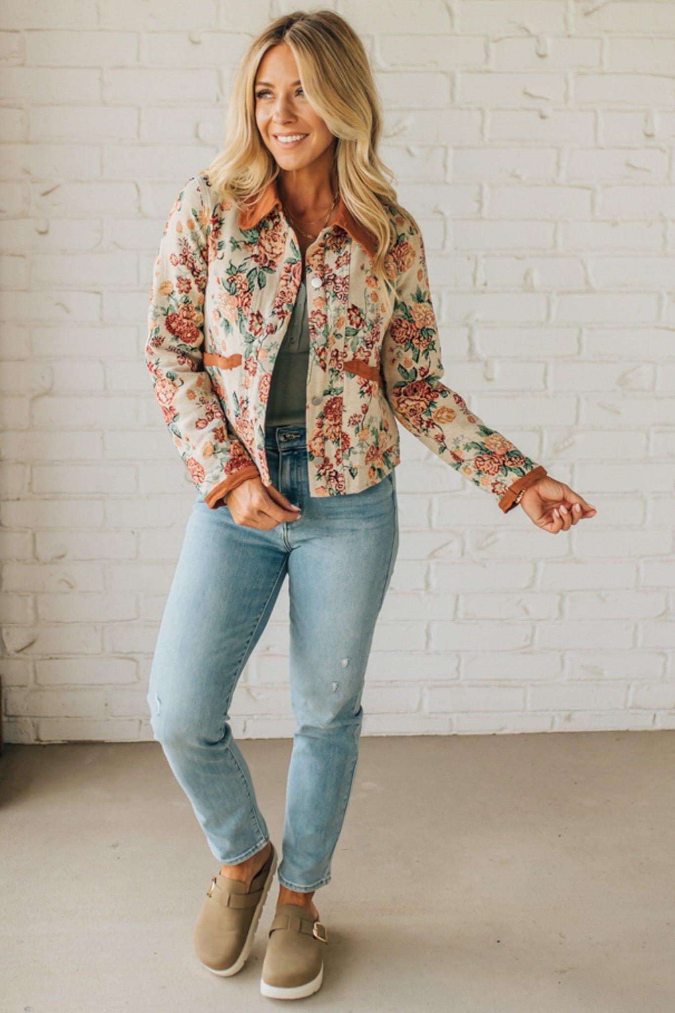 Woman wearing a floral shirt, jeans, and beige shoes against a white brick wall.