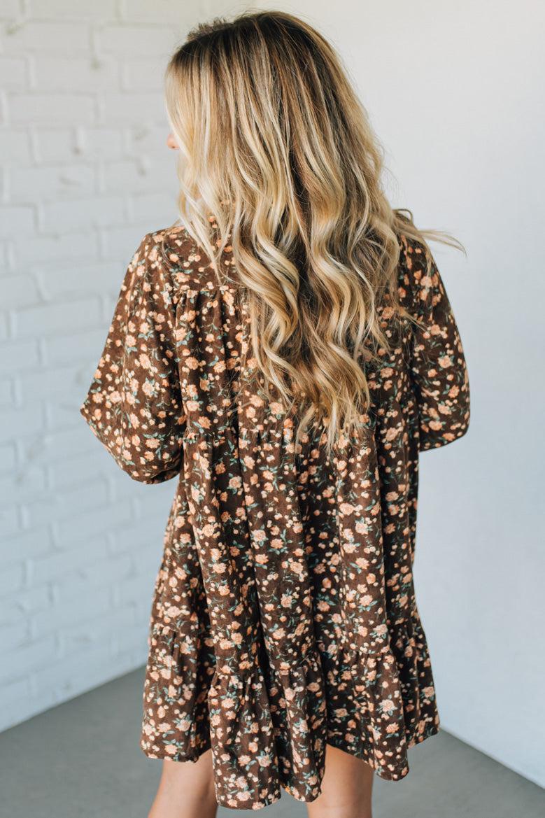 Woman wearing a brown floral dress against a white brick wall.