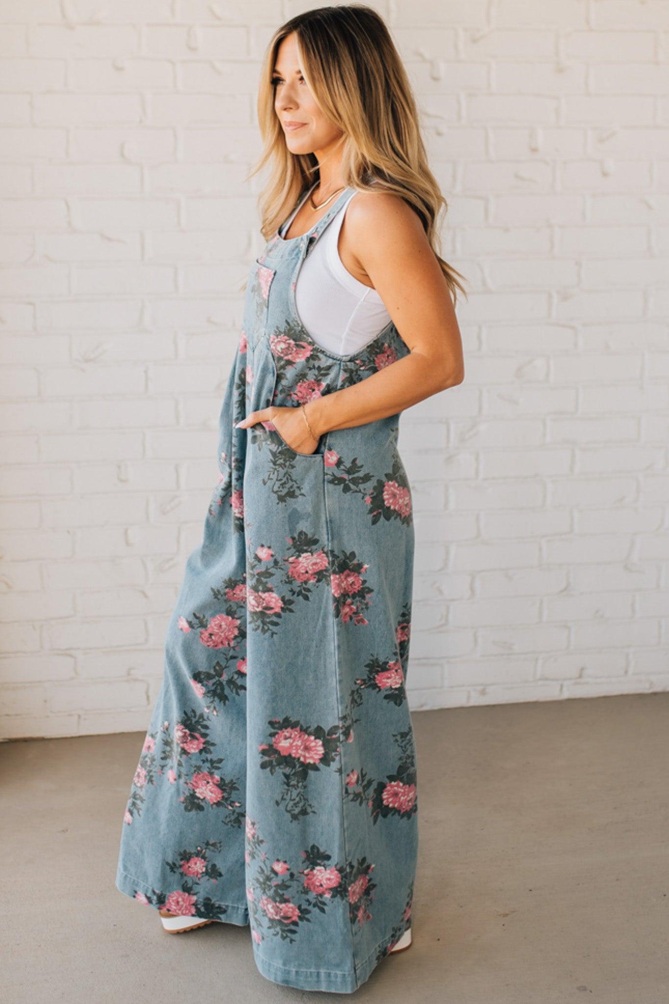 BLONDE WOMAN WEARING FLOWER PRINT WASHED DENIM WIDE LEG OVERALLS WITH ADJUSTABLE BUTTONED SHOULDER STRAP.