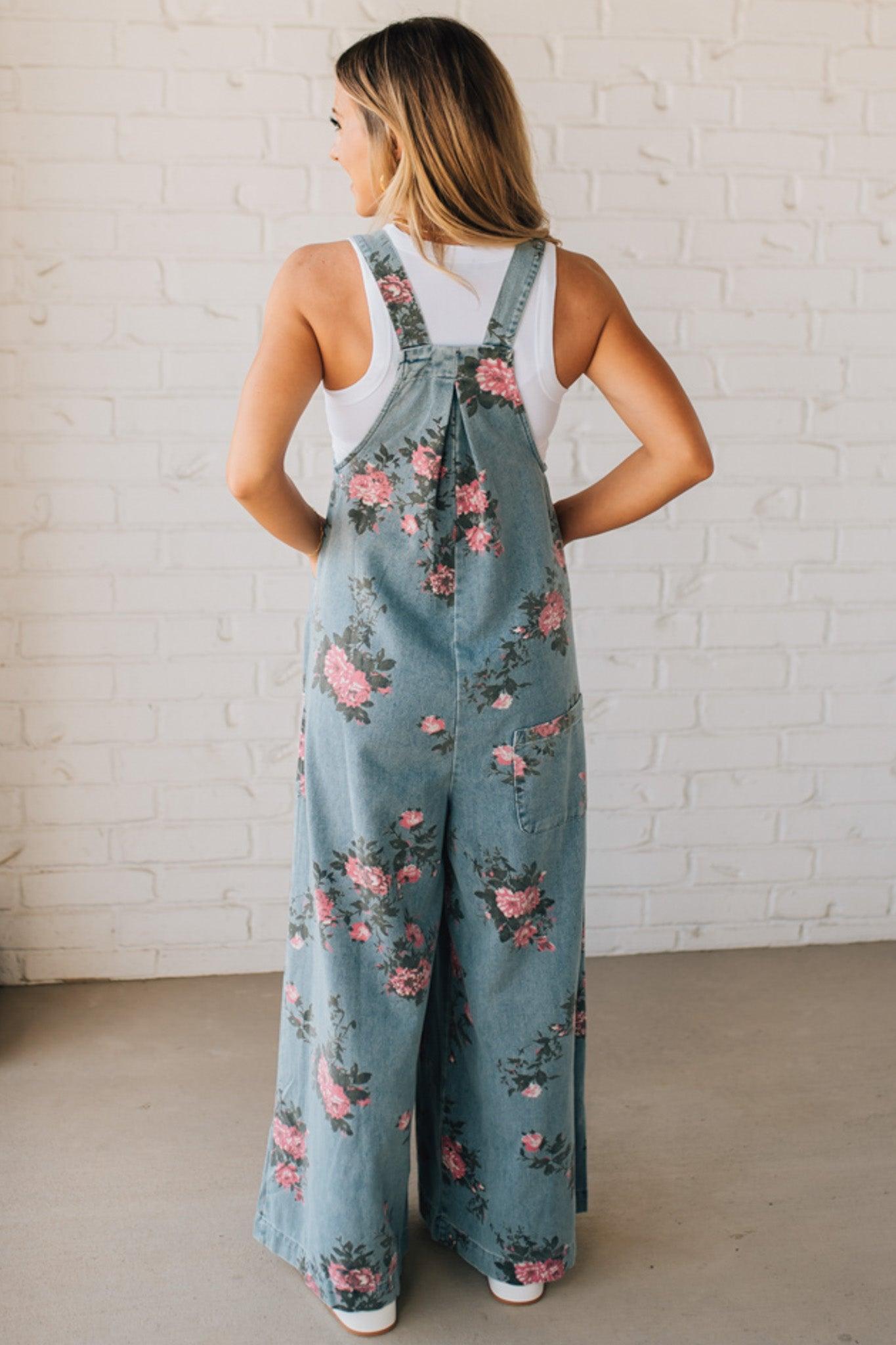 BLONDE WOMAN WEARING FLOWER PRINT WASHED DENIM WIDE LEG OVERALLS WITH ADJUSTABLE BUTTONED SHOULDER STRAP.