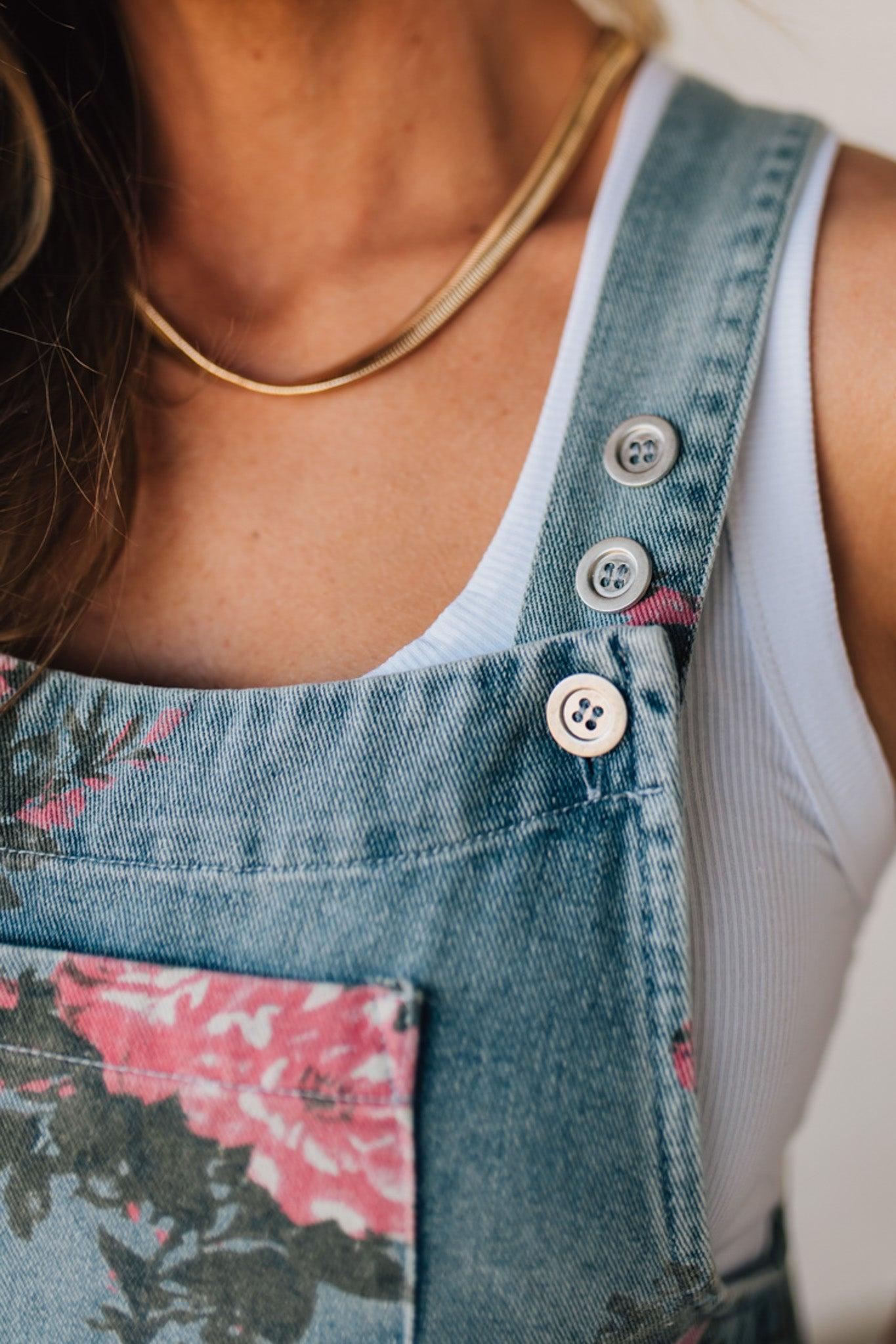 BLONDE WOMAN WEARING FLOWER PRINT WASHED DENIM WIDE LEG OVERALLS WITH ADJUSTABLE BUTTONED SHOULDER STRAP.