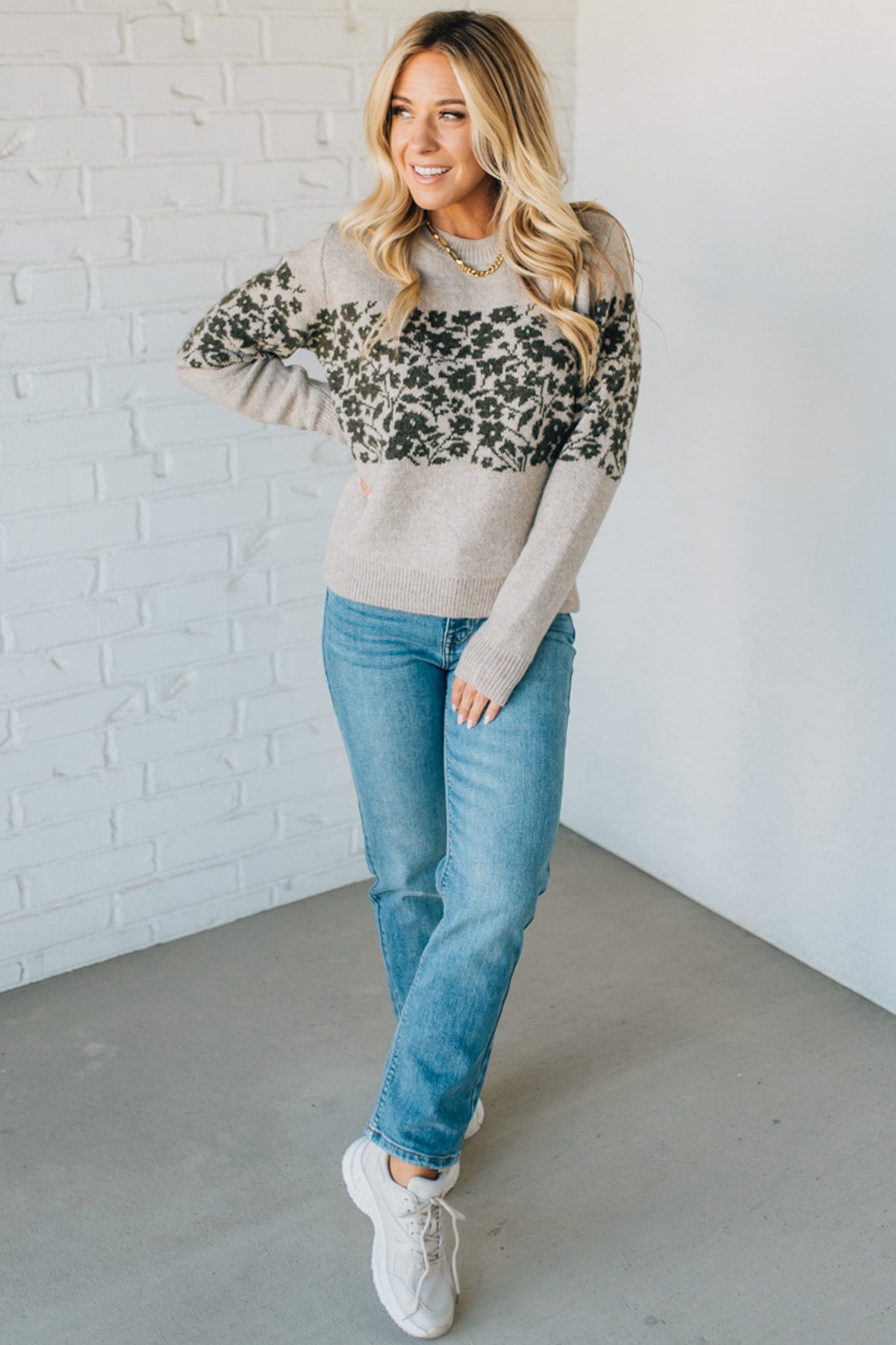Woman wearing a gray sweater with leopard print sleeves and blue jeans against a white brick wall.