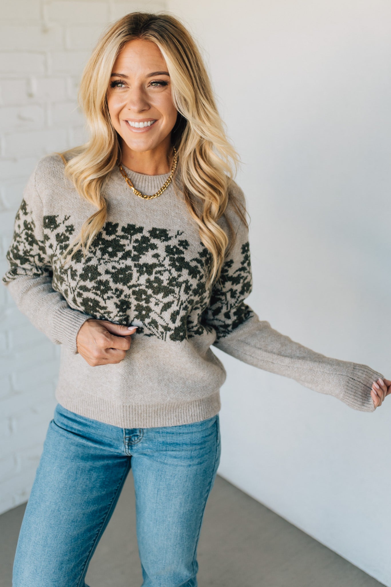 Woman wearing a gray sweater with green floral patterns and blue jeans against a white wall.