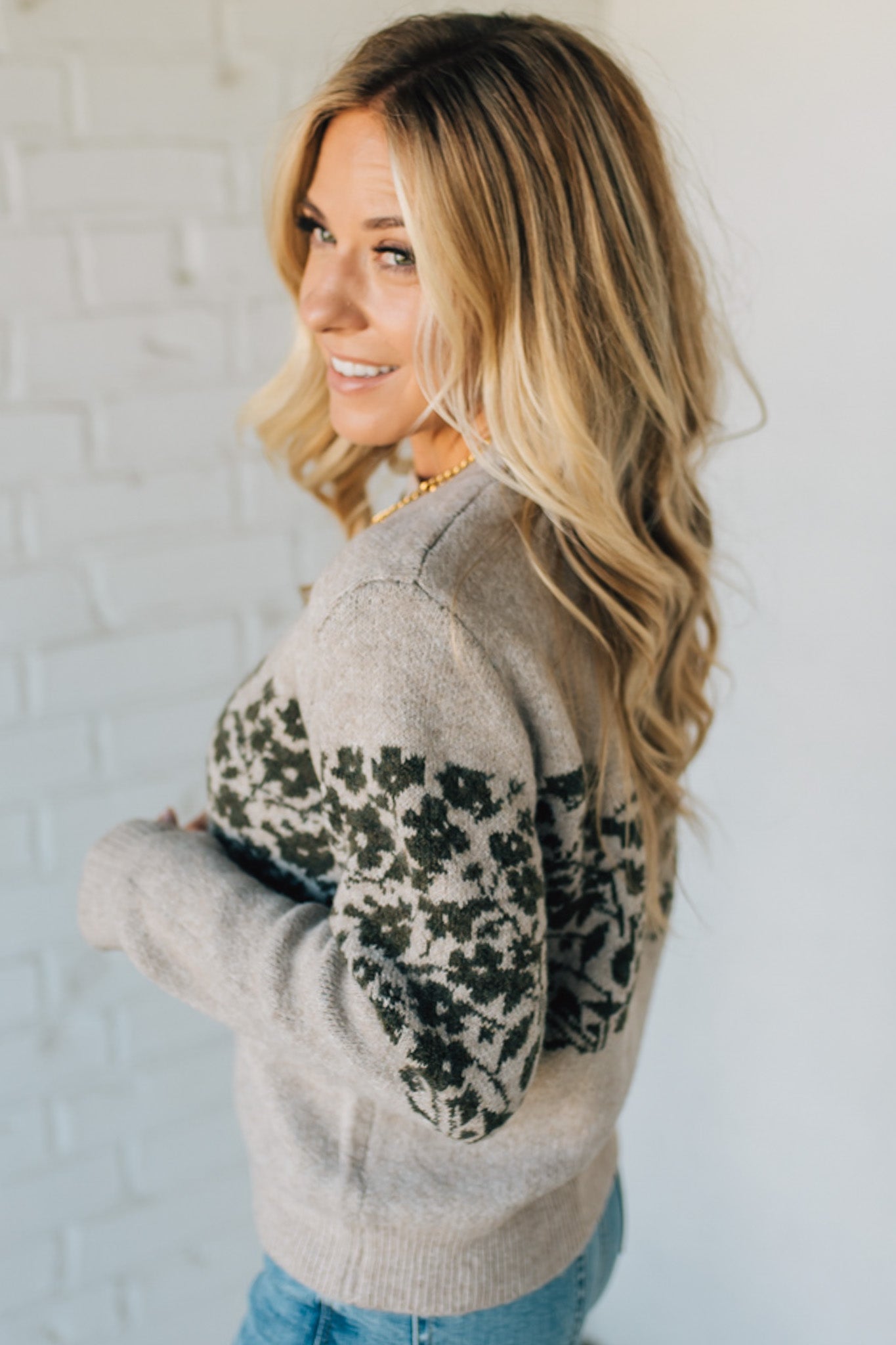 Woman wearing a gray sweater with floral patterns against a white brick wall.