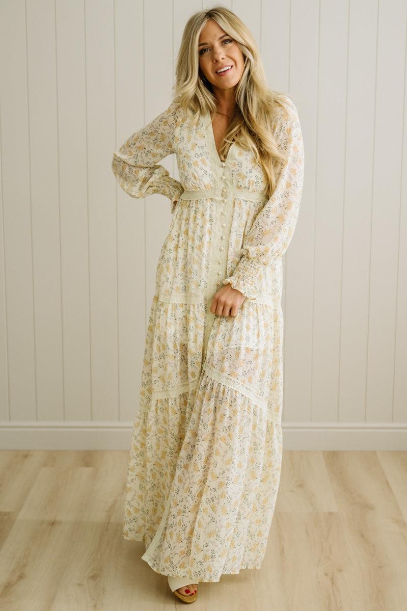 Woman wearing a long floral dress standing against a white paneled wall.