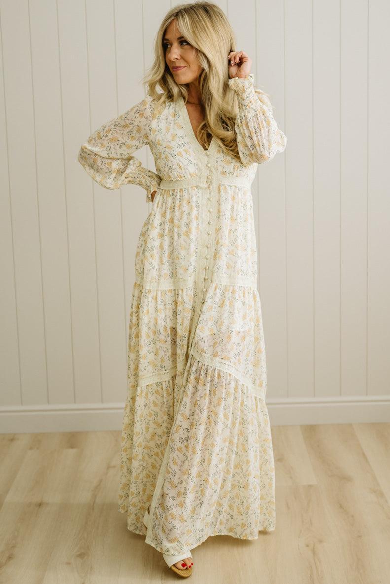 Woman wearing a long floral dress against a white paneled wall.