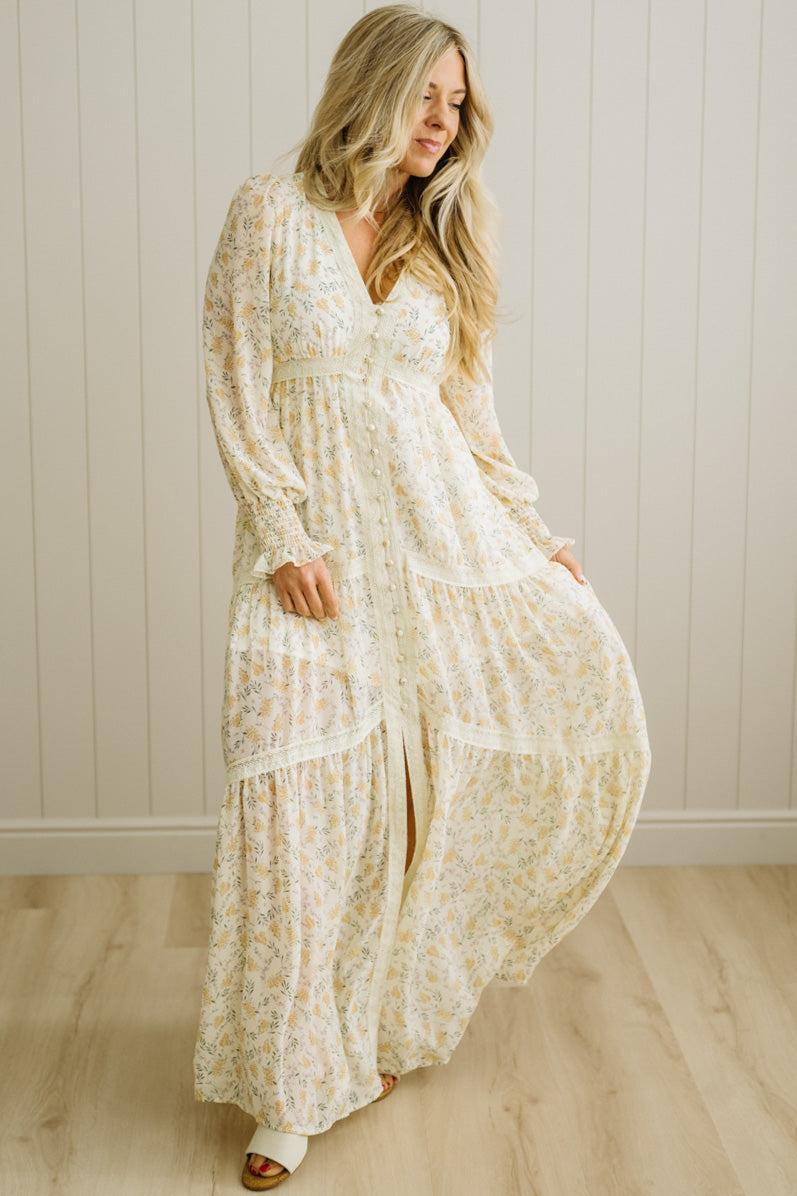 Woman wearing a long, light-colored floral dress against a white paneled wall.