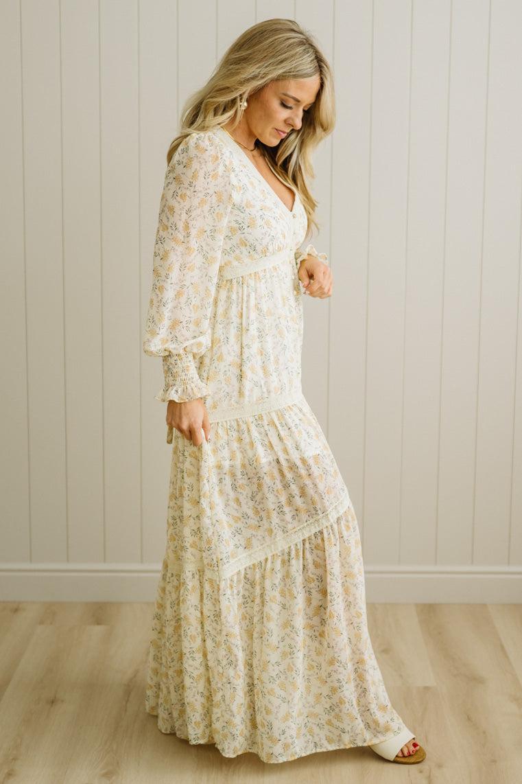 Woman wearing a long floral dress against a white paneled wall.