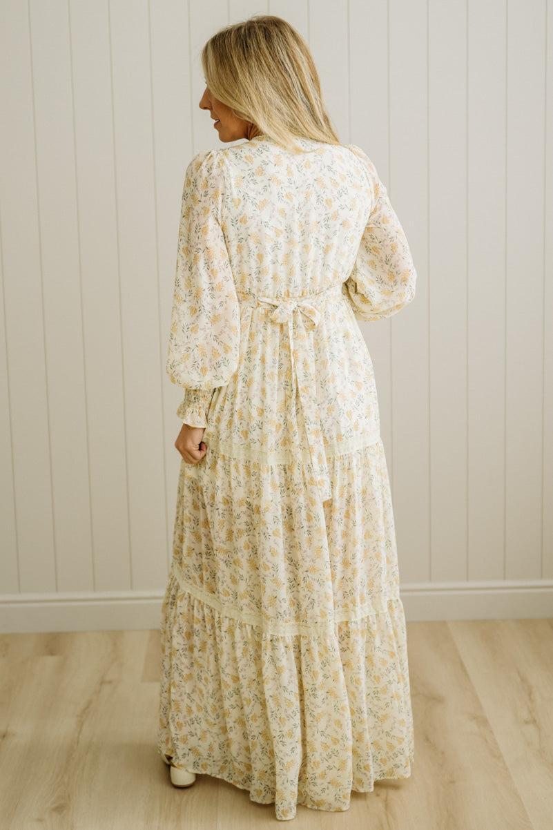 Woman wearing a long floral dress against a white paneled wall.