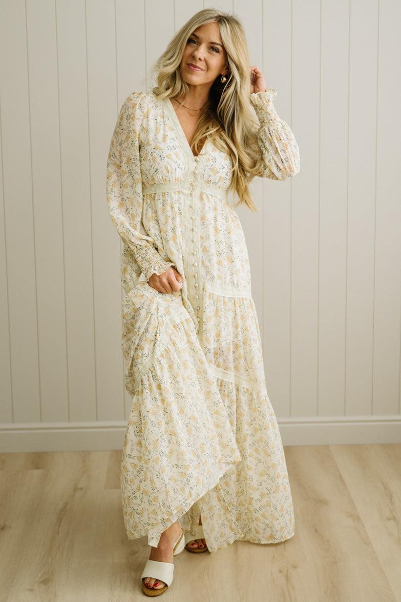 Woman wearing a long, light-colored floral dress against a white paneled wall.