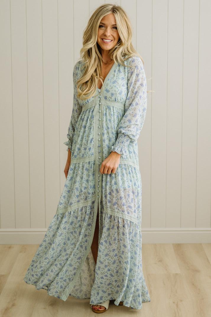 Woman wearing a long floral dress against a white paneled wall.