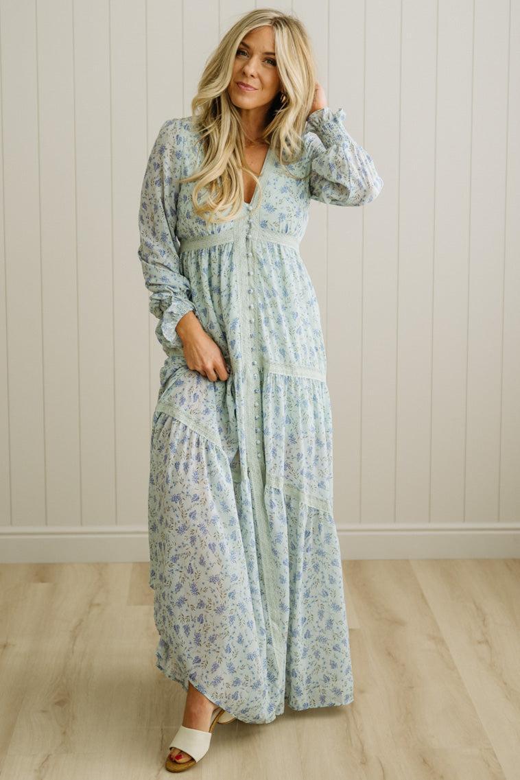 Woman wearing a floral dress standing against a light wooden paneled wall.