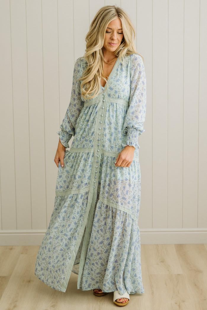 Woman wearing a long floral dress standing against a white paneled wall.