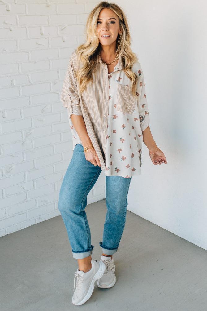 Woman wearing a patterned shirt, blue jeans, and white sneakers against a white brick wall.