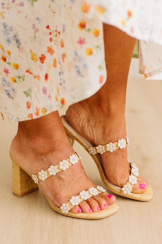 Beige sandals with floral embellishments worn with a floral dress on a neutral background