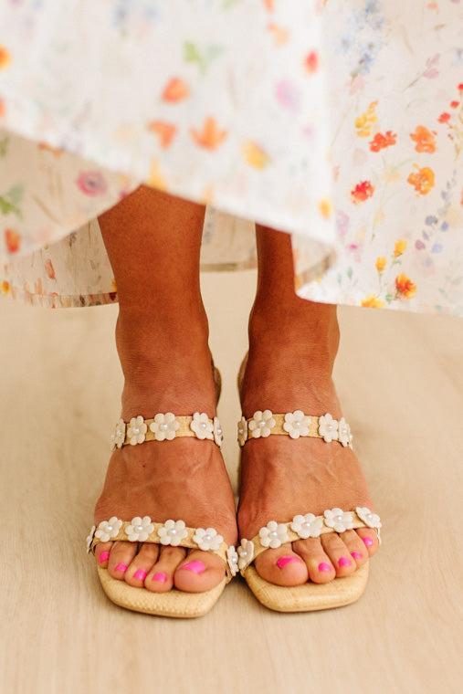 Brown sandals with floral embellishments worn by a person in a floral dress.