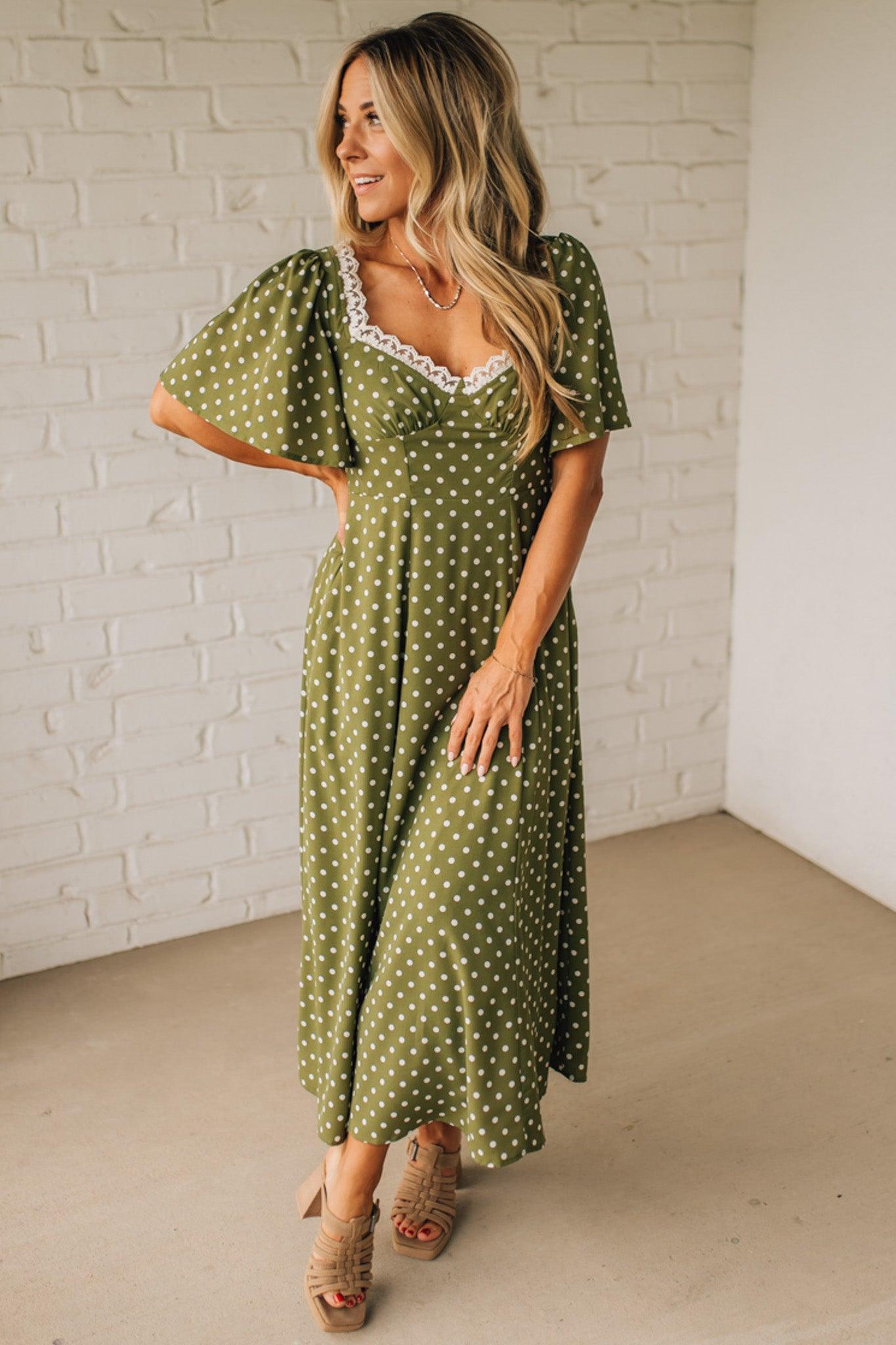 Woman wearing a green polka dot dress with lace trim against a white brick wall.