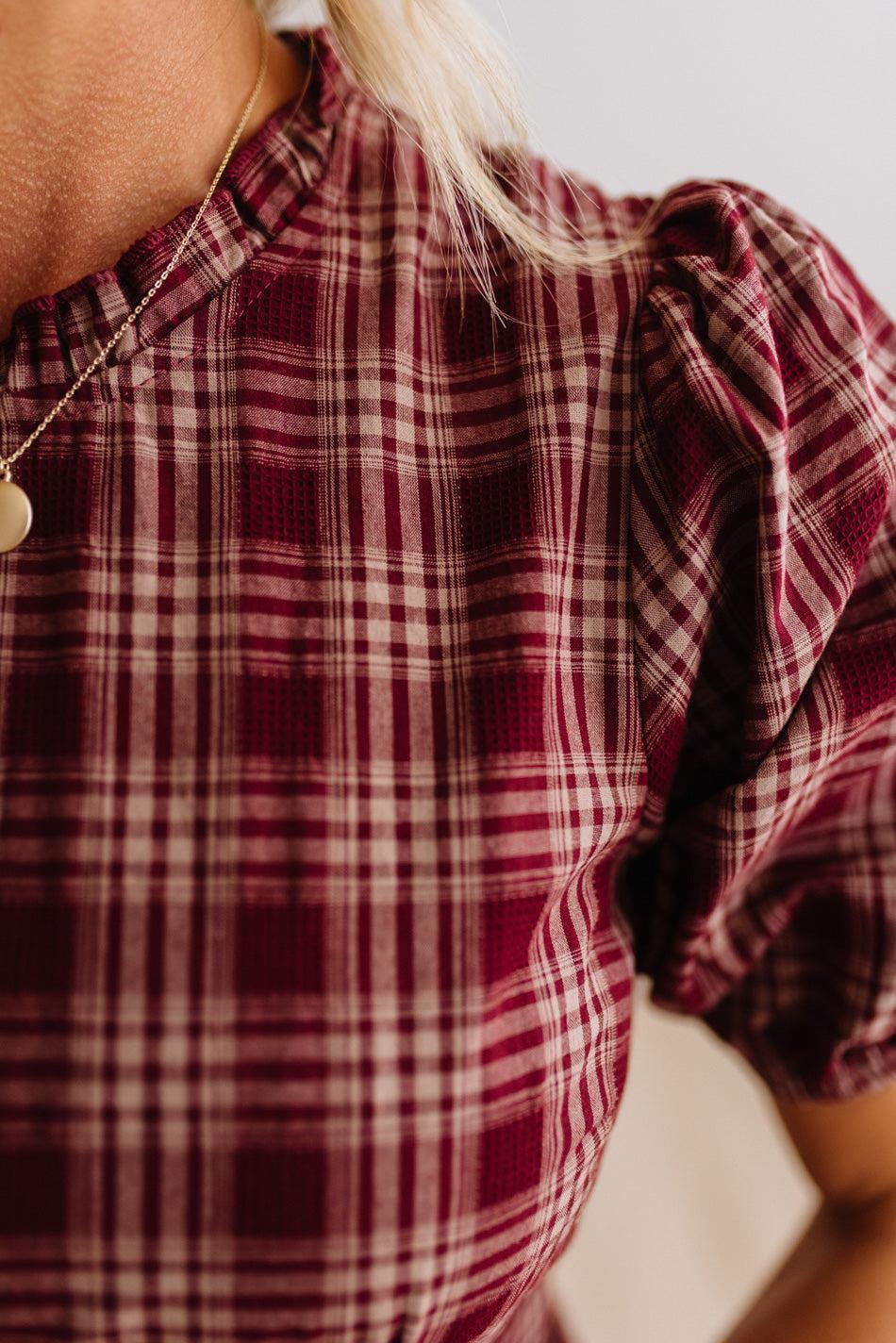 Close-up of a person wearing a burgundy and cream plaid top with puff sleeves and a high ruffled neckline, paired with a gold necklace.