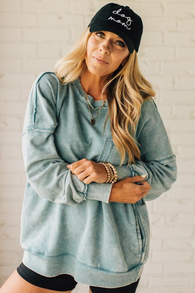 Blonde woman wearing a boyfriend fit drop shoulder sweatshirt pullover, with front pocket accent, exposed raw seams, and all over acid wash detail.