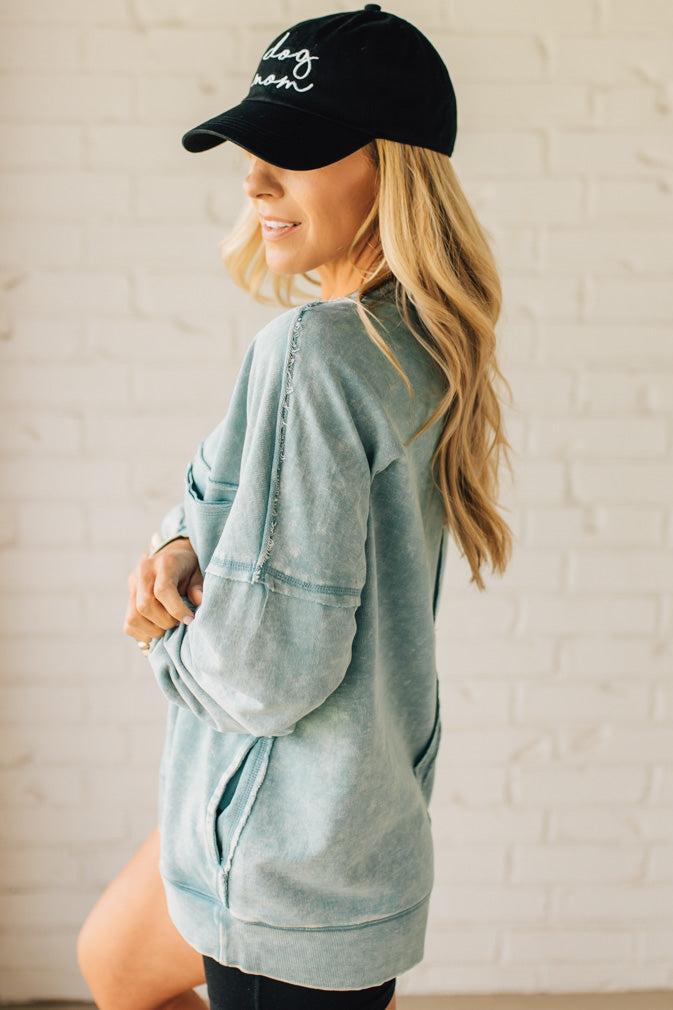 Blonde woman wearing a boyfriend fit drop shoulder sweatshirt pullover, with front pocket accent, exposed raw seams, and all over acid wash detail.