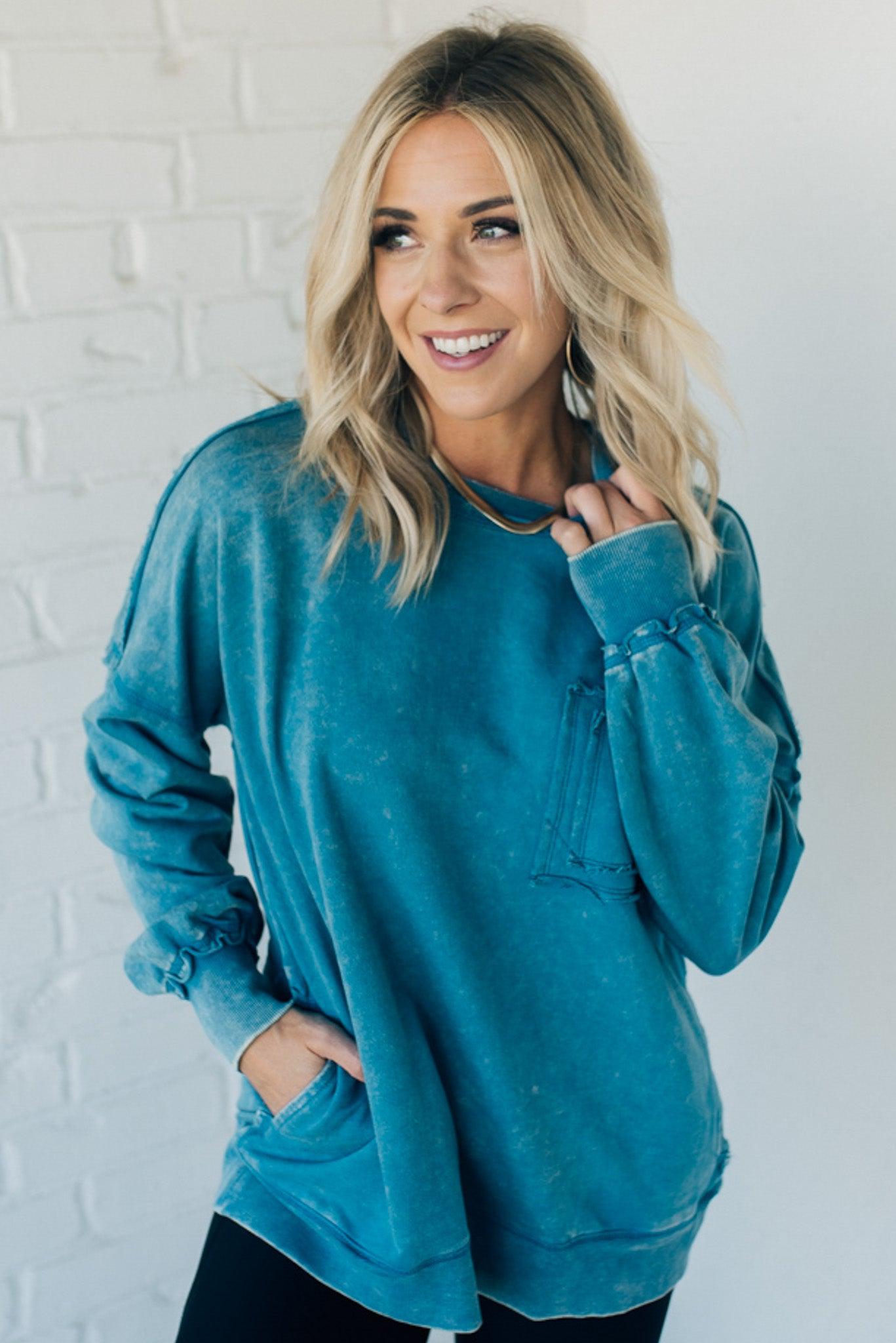 Blonde woman wearing a boyfriend fit drop shoulder sweatshirt pullover, with front pocket accent, exposed raw seams, and all over acid wash detail.
