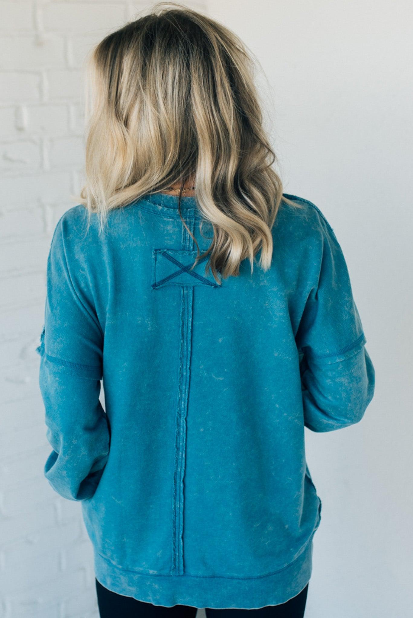 Blonde woman wearing a boyfriend fit drop shoulder sweatshirt pullover, with front pocket accent, exposed raw seams, and all over acid wash detail.