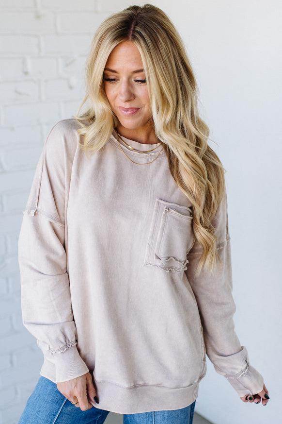 Woman wearing a beige sweatshirt with a pocket against a white brick wall.