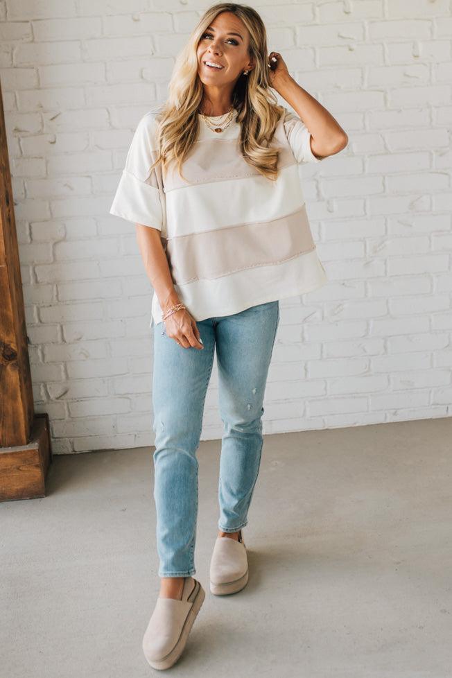 Woman wearing a white layered top and light blue jeans against a white brick wall.