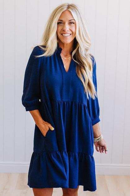 Blonde woman wearing a knee length solid dress with tiered skirt, v-neckline and side pockets in navy.