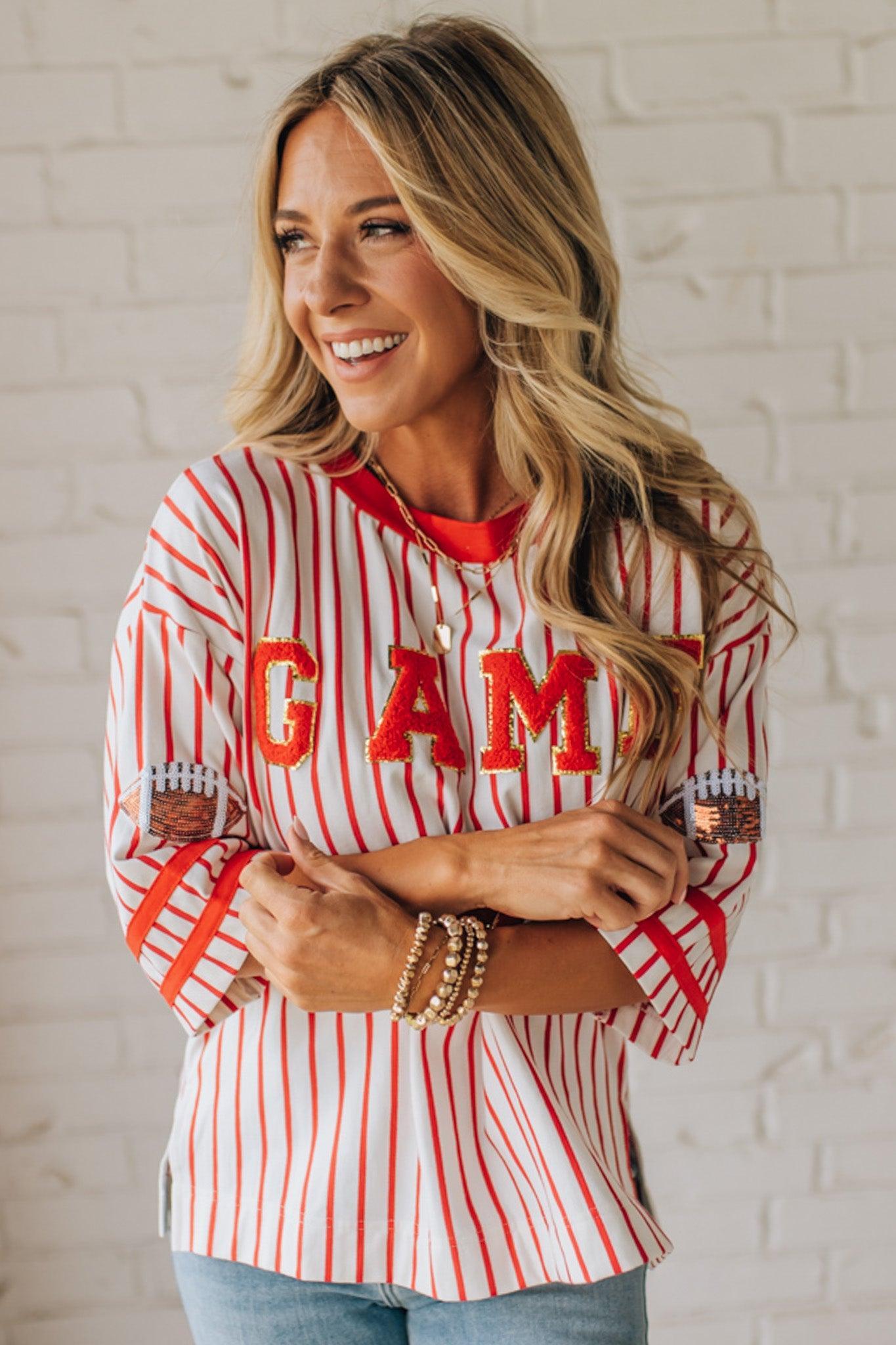 Woman wearing a red and white striped shirt with 'GAME DAY' text, standing against a white brick wall.