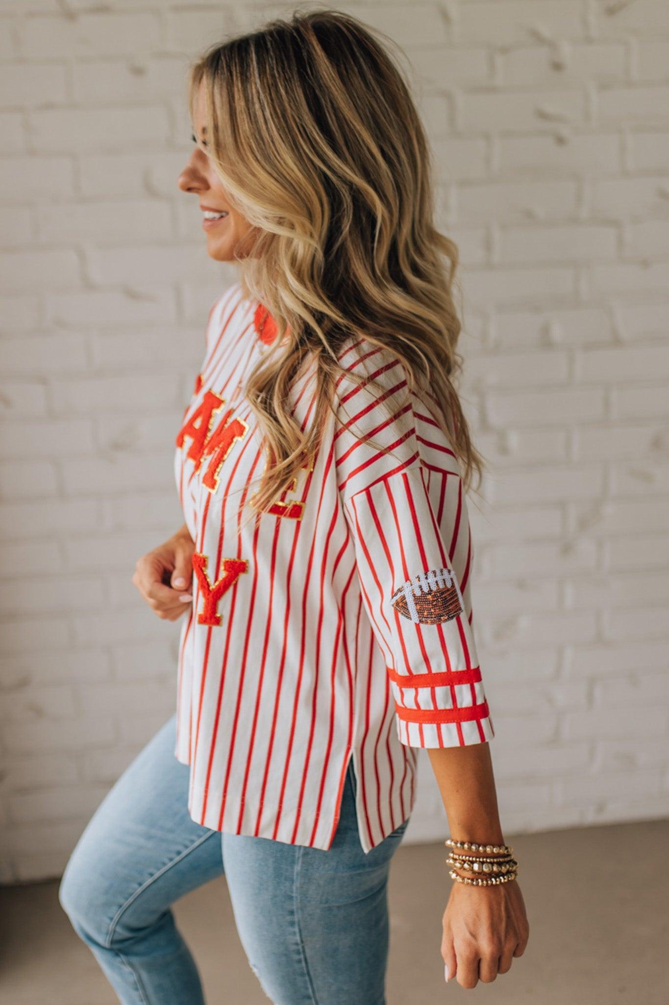 Woman wearing a red and white striped shirt with 'GAME DAY' text, standing against a white brick wall.
