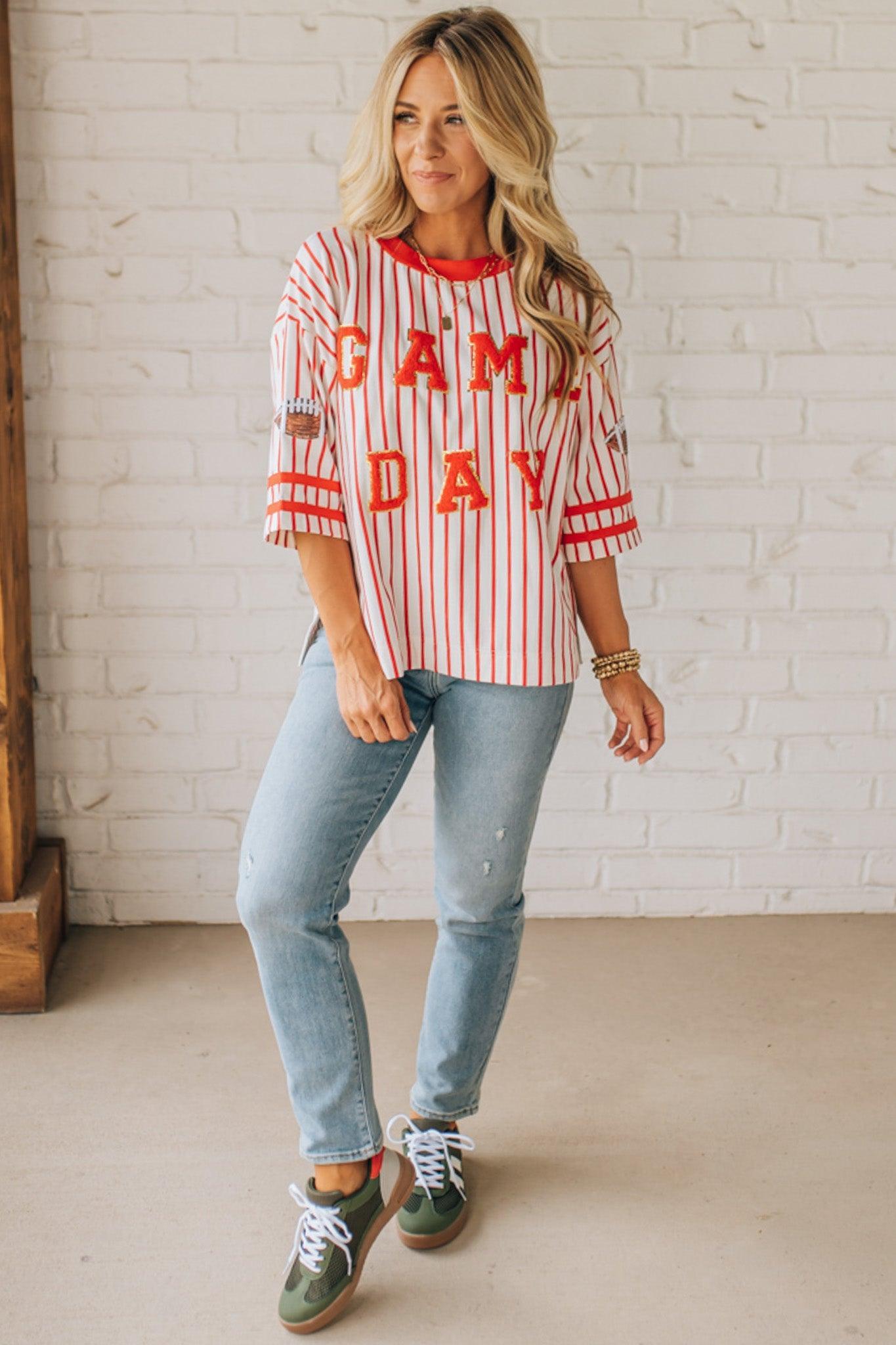 Woman wearing a 'Game Day' striped shirt with light blue jeans against a white brick wall.