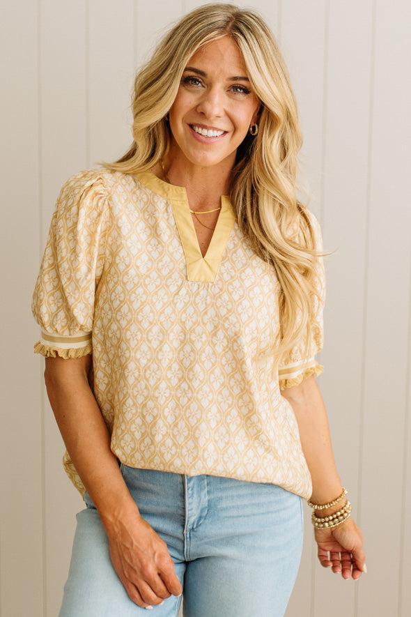 Woman wearing a yellow patterned blouse and light blue jeans against a neutral background