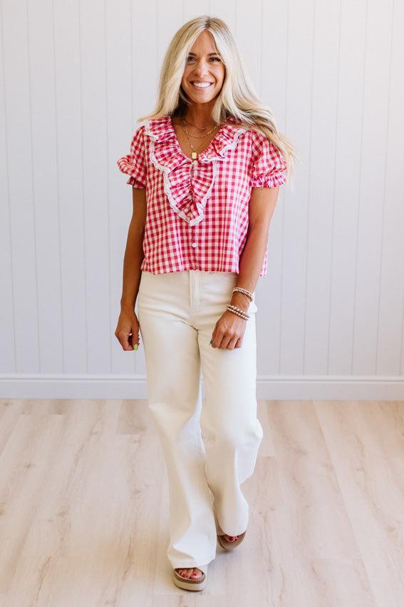 Woman wearing a red and white checkered blouse with ruffled sleeves and white pants indoors.