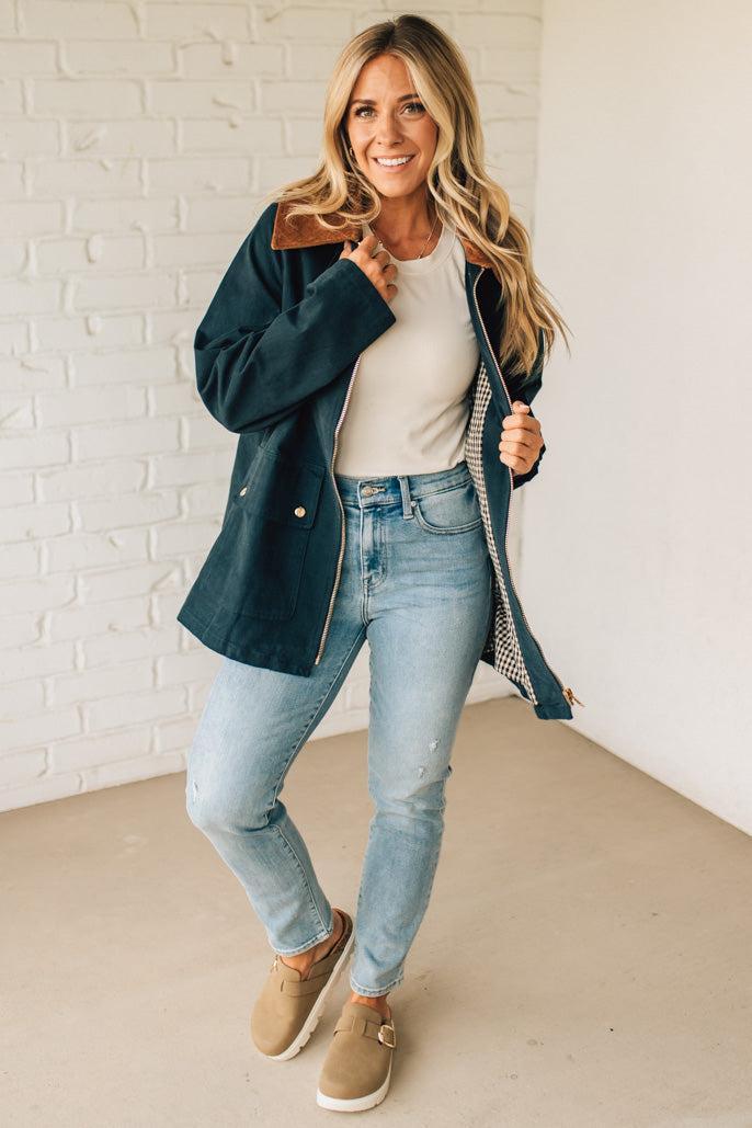 Woman wearing a dark blue jacket, white shirt, light blue jeans, and beige boots against a white brick wall.
