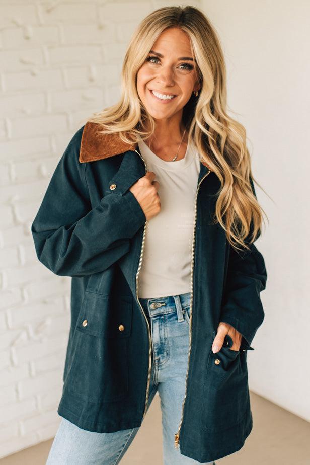 Woman wearing a dark blue coat with brown leather accents against a white brick wall.
